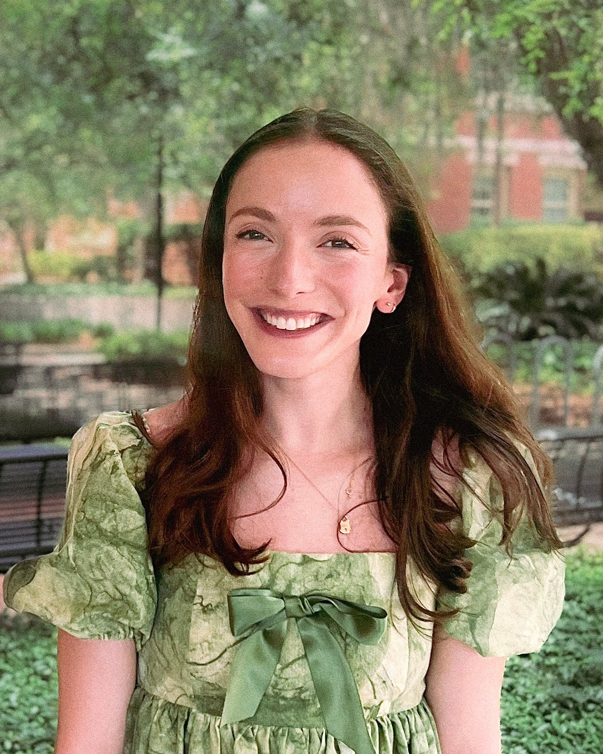 A headshot of a young woman with long brunette hair. She is smiling, looking directly at the camera. She wears a green dress which matches her surroundings of landscaping and trees.